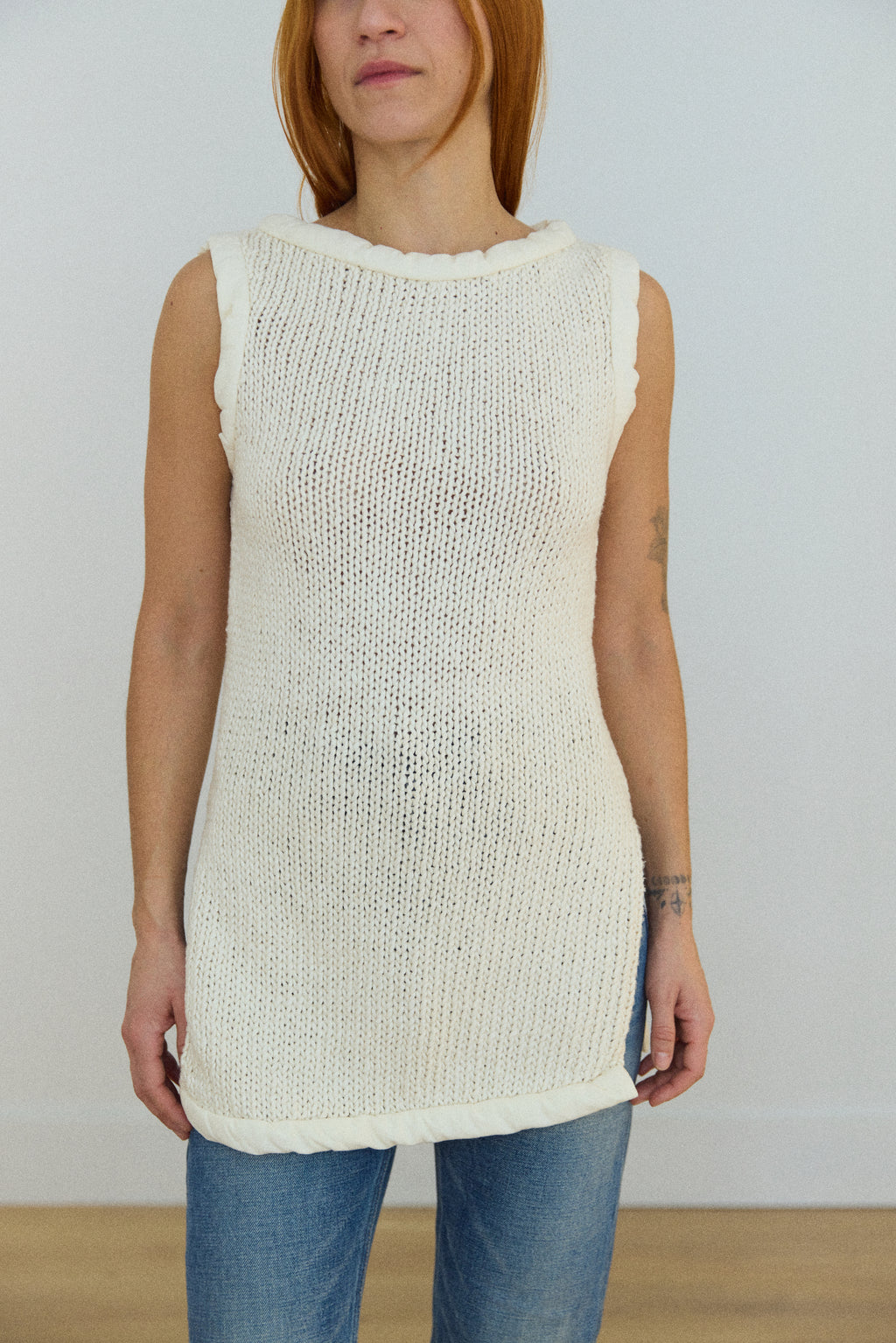 Woman wearing a sleeveless cream knit top and blue jeans against a plain background