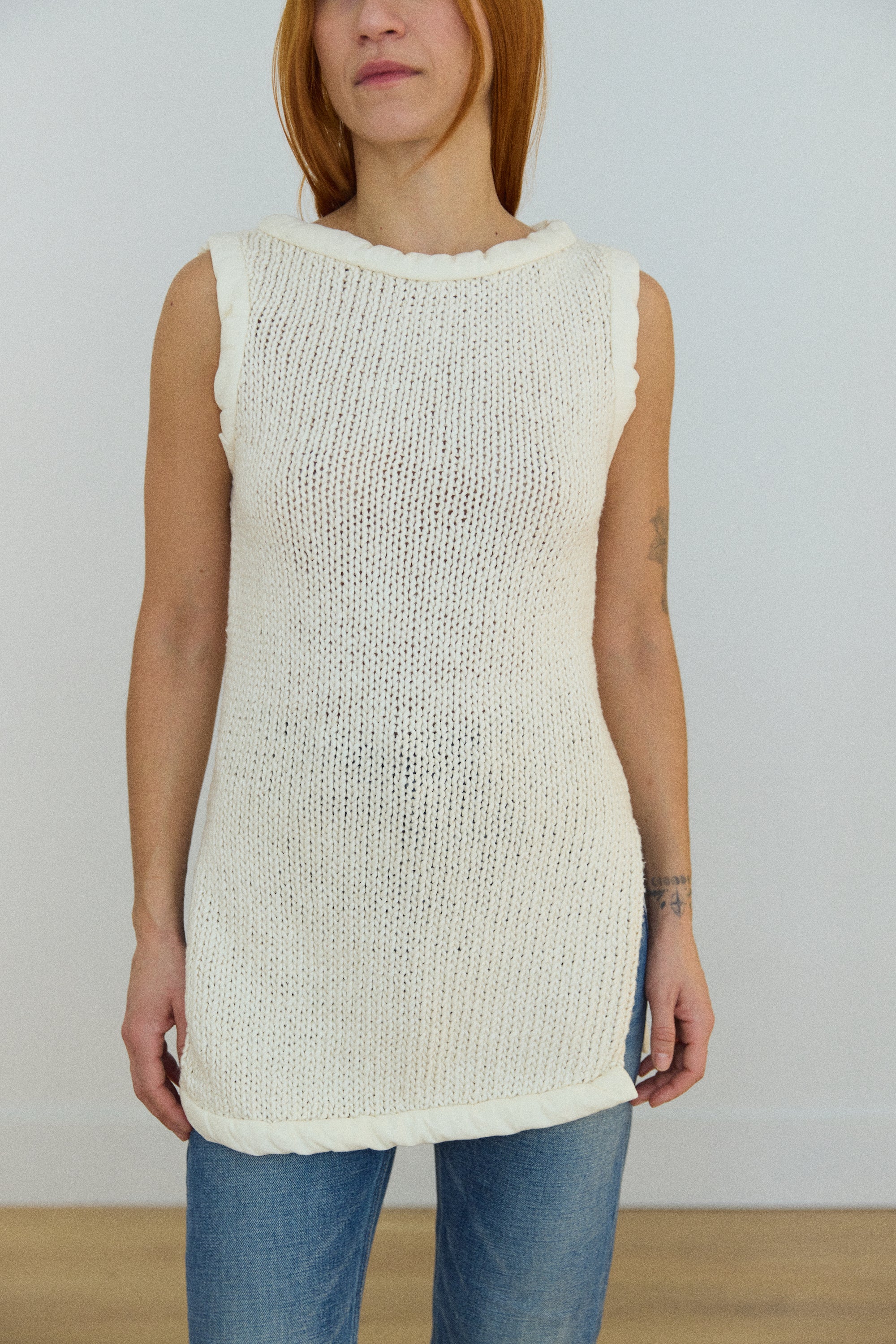Woman wearing a sleeveless cream knit top and blue jeans against a plain background