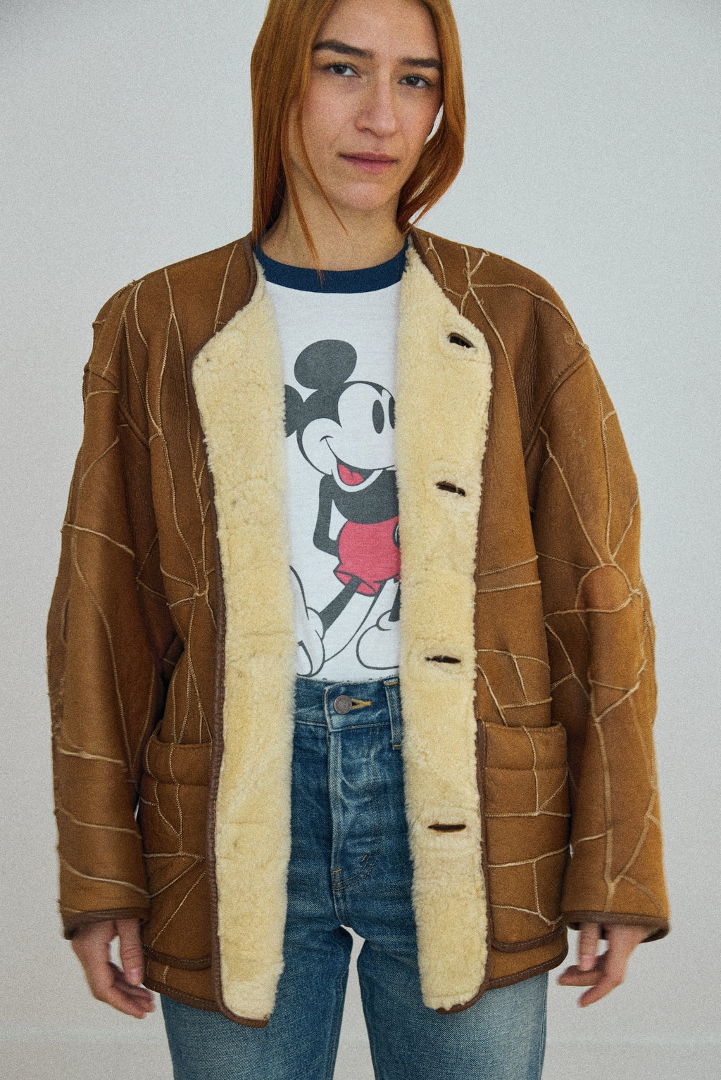Person wearing a brown shearling jacket over a Mickey Mouse t-shirt on a plain background