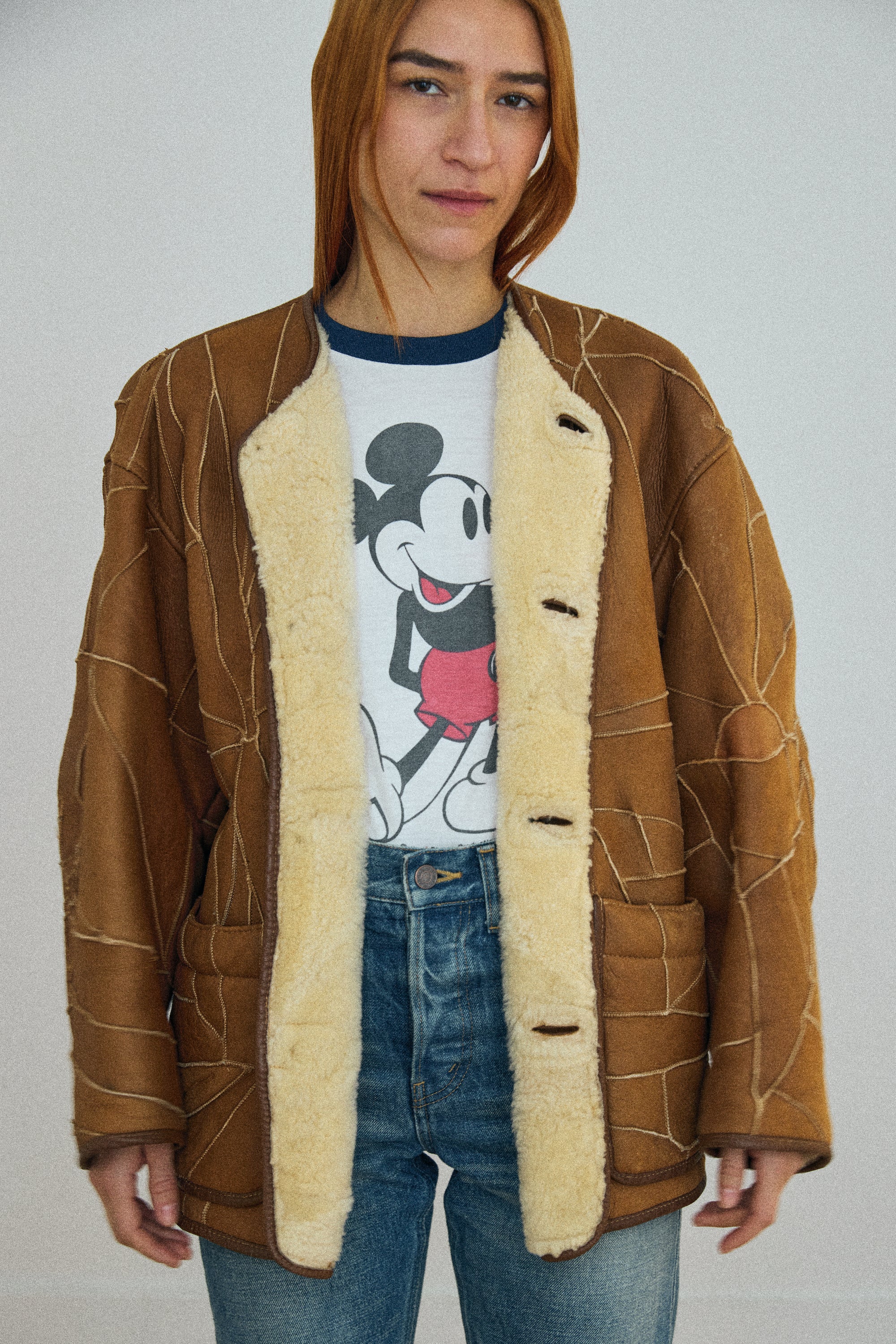 Person wearing a brown shearling jacket over a Mickey Mouse t-shirt on a plain background