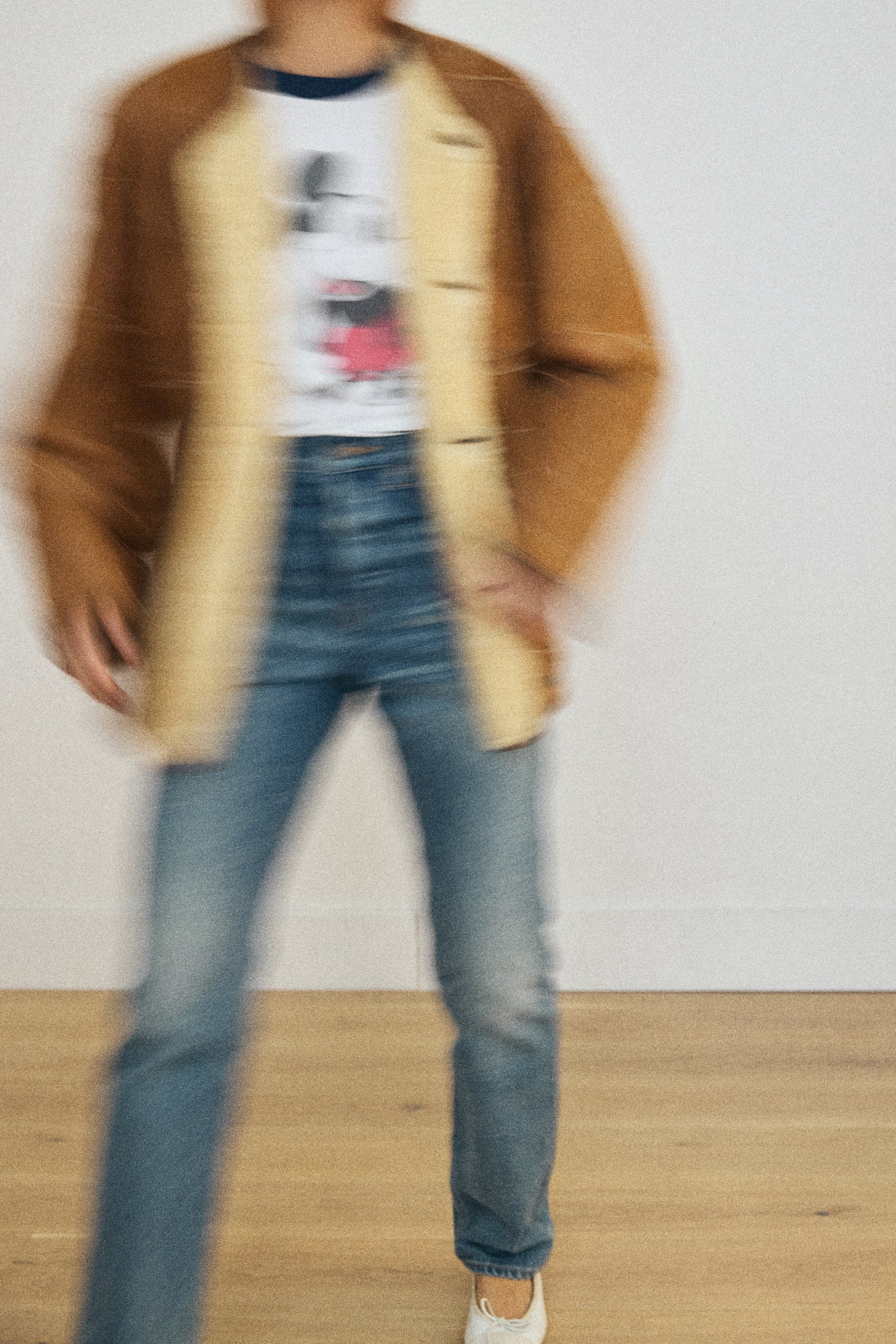 Person wearing a brown coat, graphic t-shirt, and blue jeans on a plain background