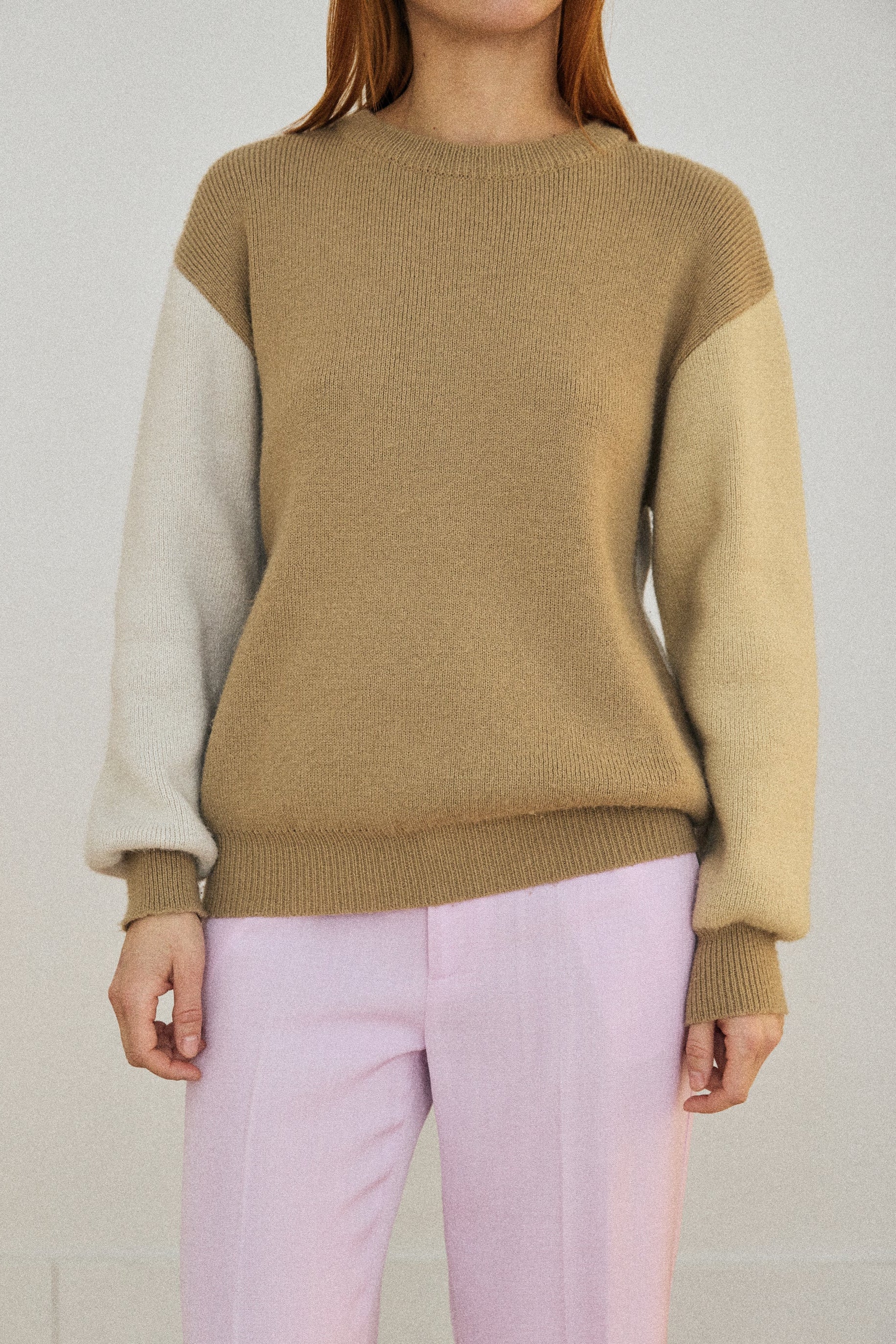 Person wearing a beige sweater with color-blocked sleeves and light pink pants against a plain background