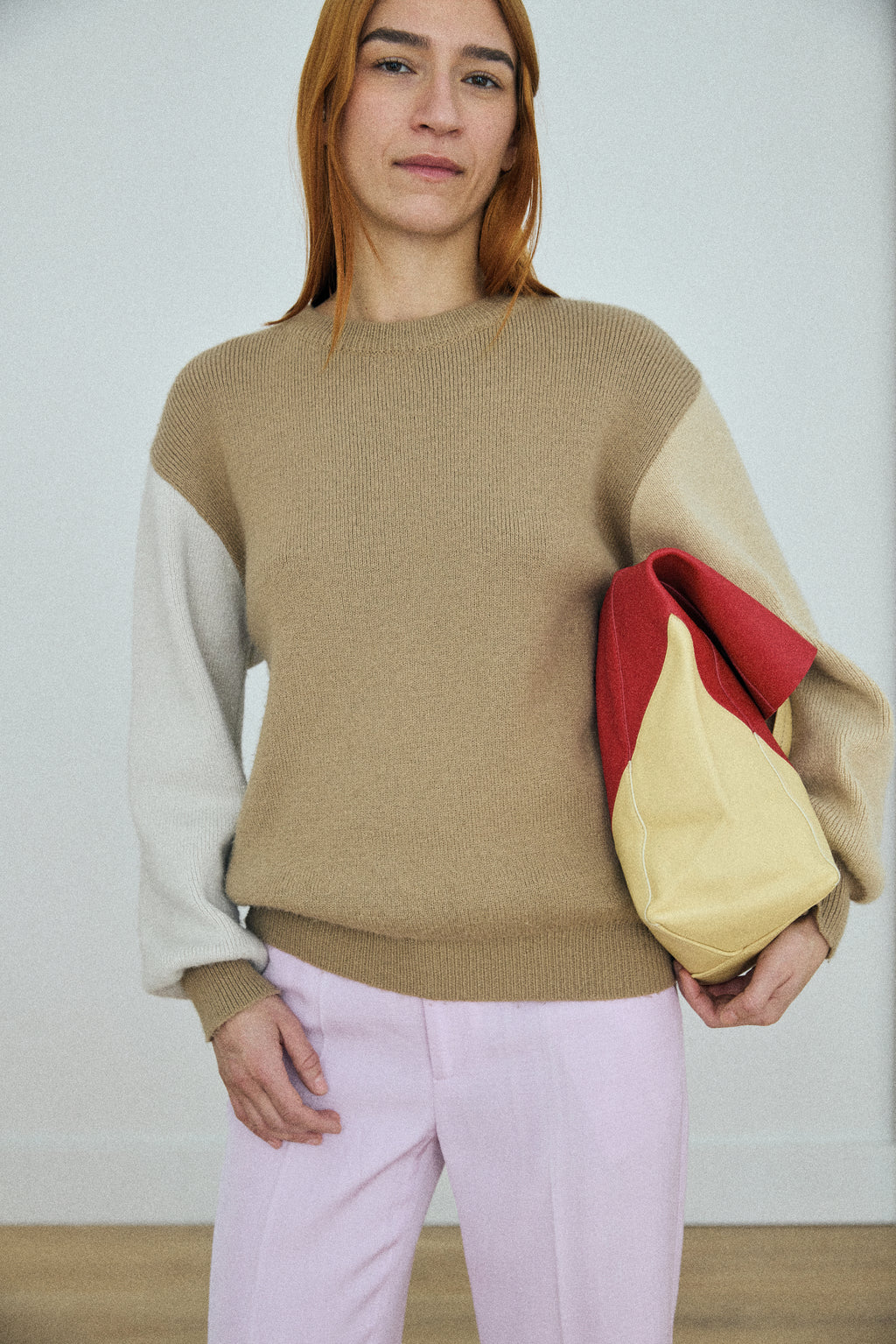 Person wearing a color-blocked sweater holding a red and beige bag against a white background