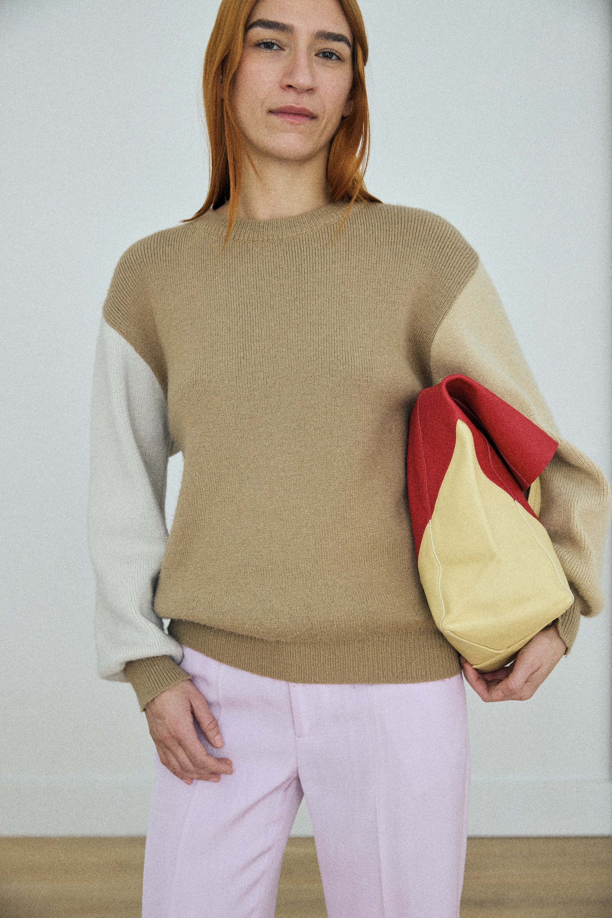 Person wearing a color-blocked sweater holding a red and beige bag against a white background