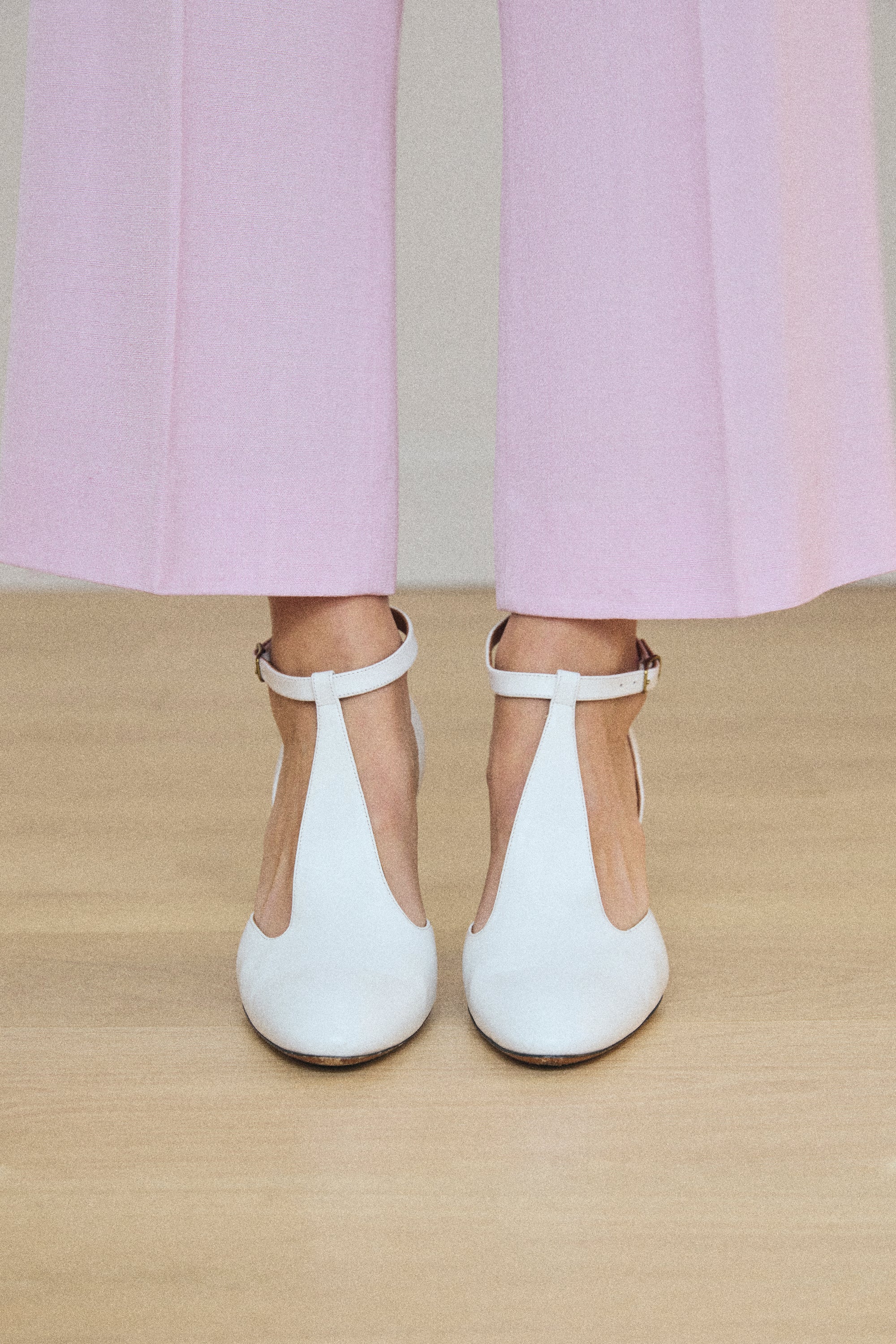 White shoes with thin straps worn with light pink pants on a beige floor.