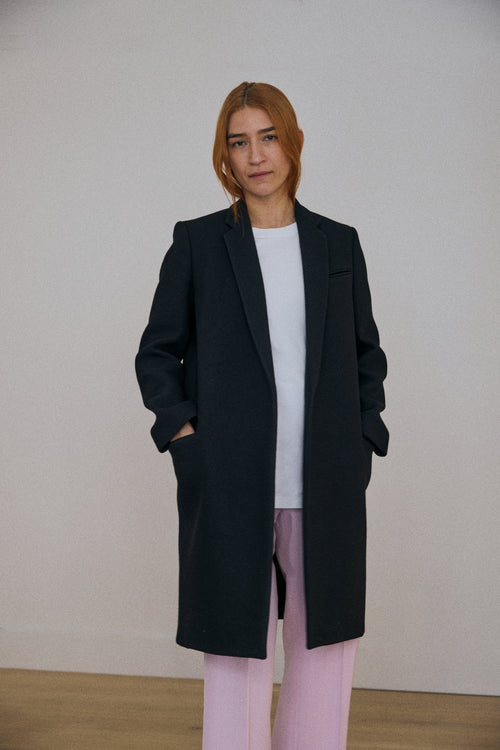 Person wearing a black coat, white shirt, and pink pants against a plain background