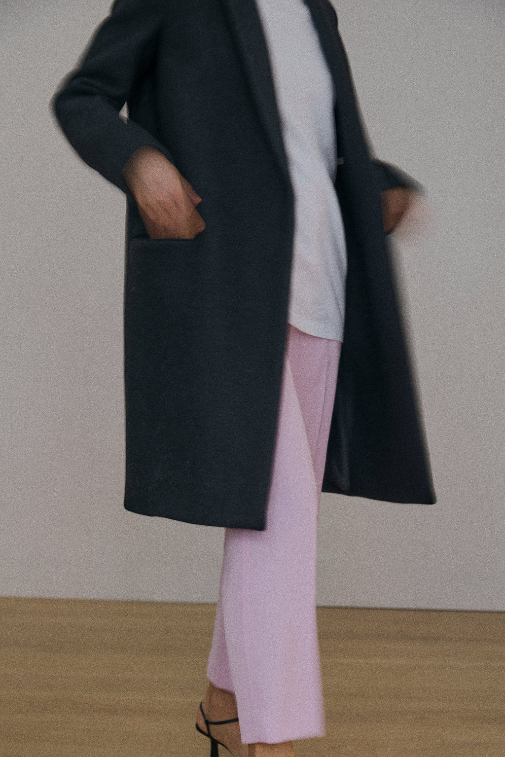 Person wearing a long dark coat over a light shirt, pink pants, and black high-heeled sandals on a plain background.
