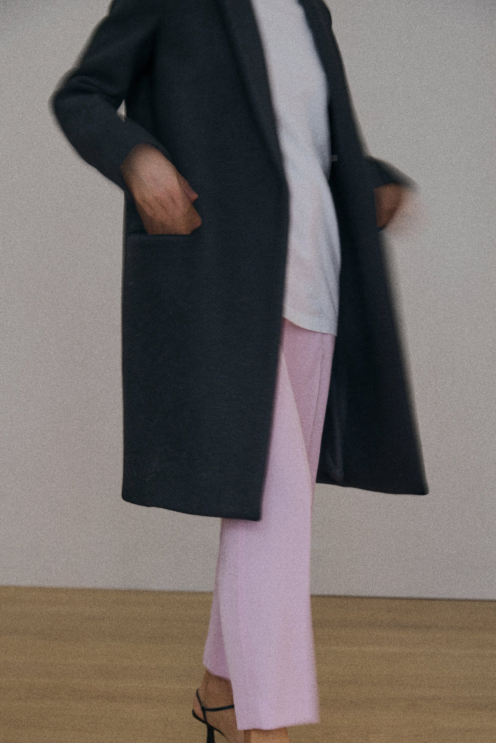 Person wearing a long dark coat over a light shirt, pink pants, and black high-heeled sandals on a plain background.
