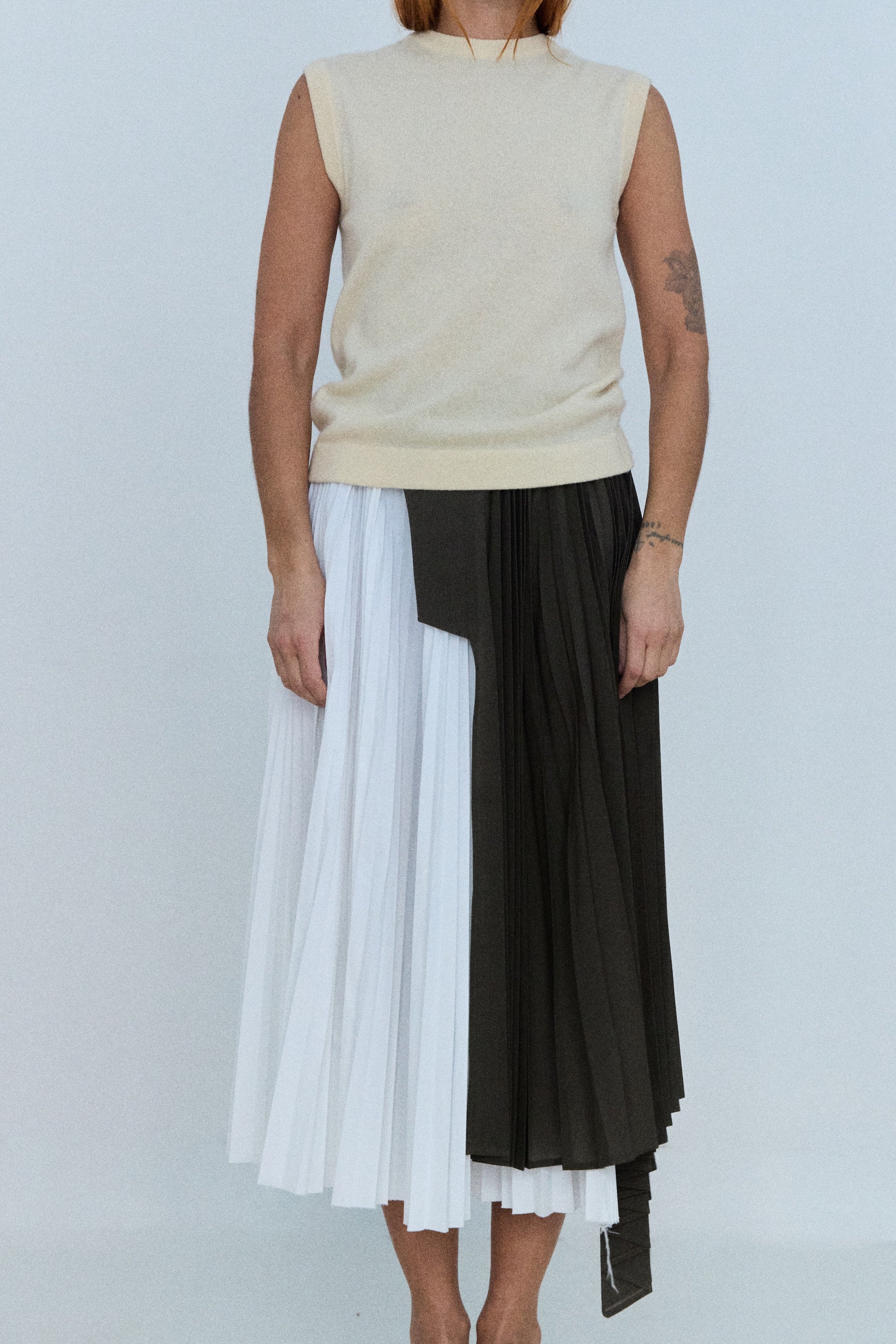Person wearing a beige sleeveless top and a black and white pleated skirt on a plain background
