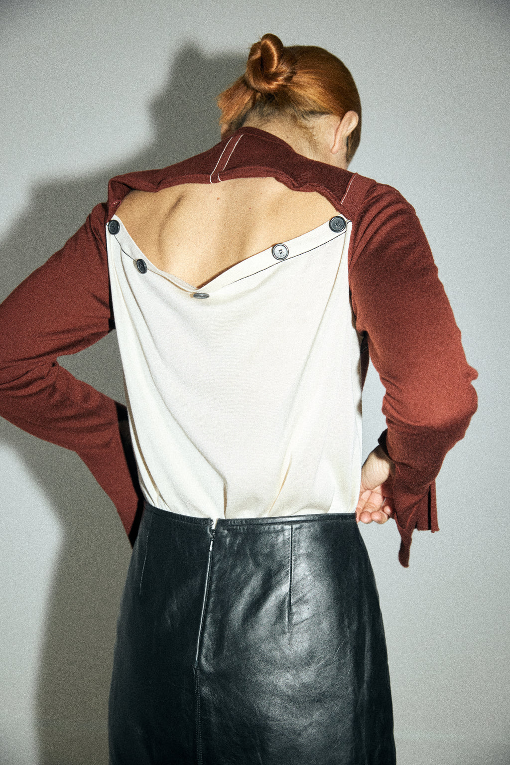 Person wearing a unique outfit with a white top, red cardigan, and black leather skirt against a plain background.