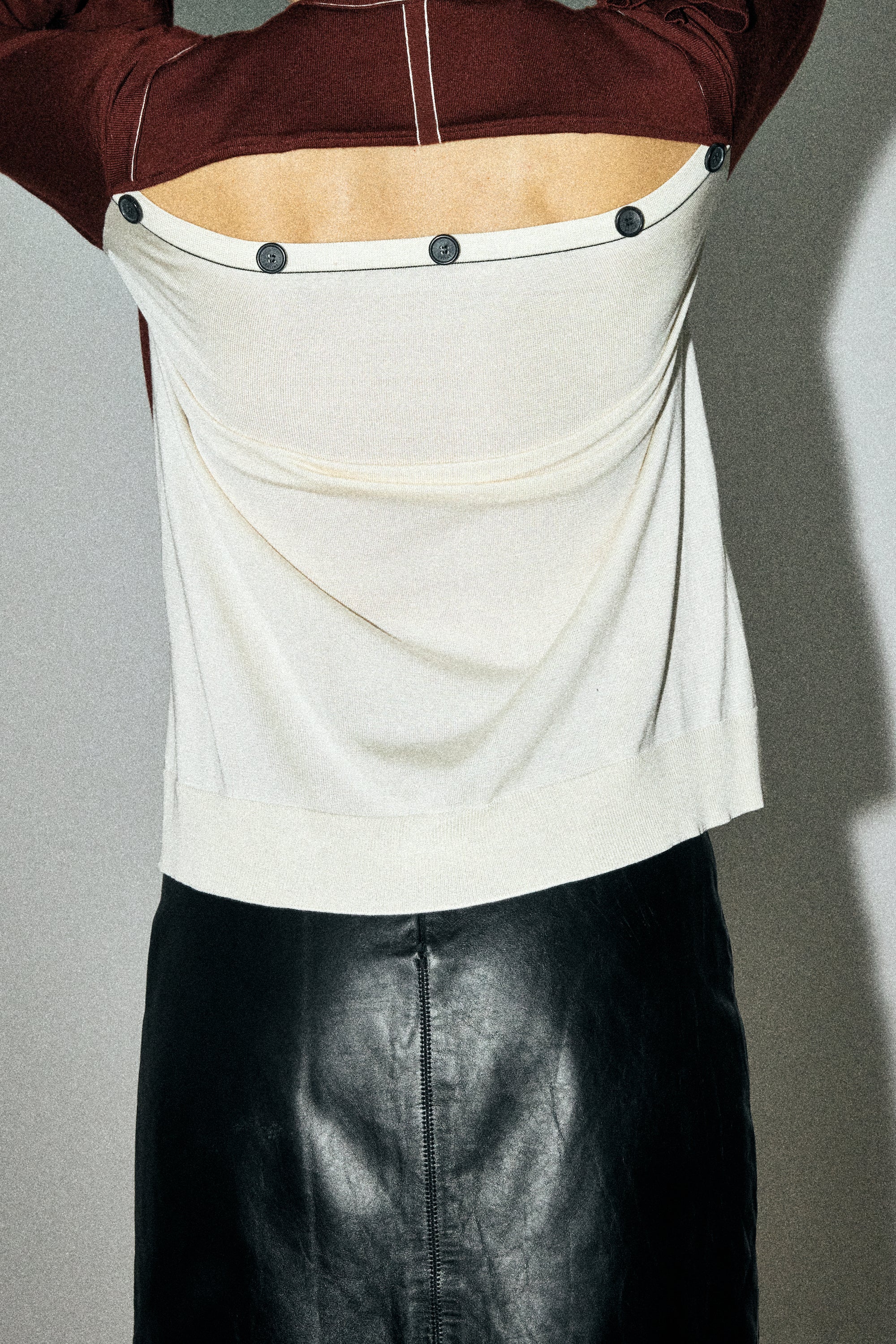 Person wearing a white top with black buttons and a brown jacket over it, standing against a gray background.