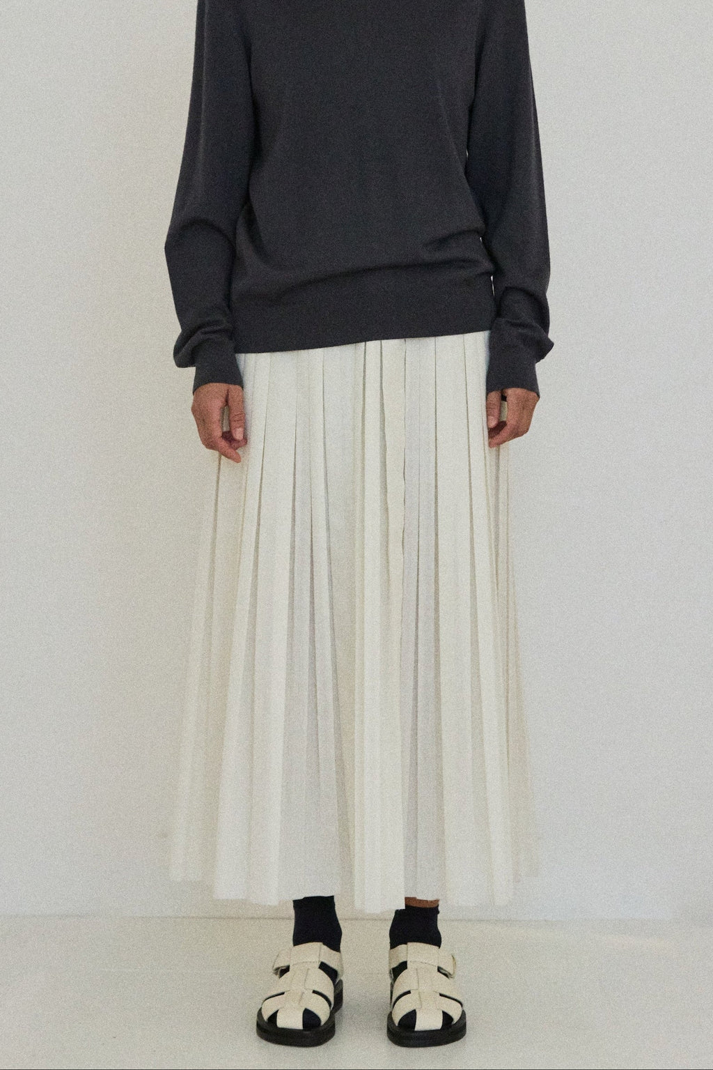 Person wearing a dark turtleneck sweater and white pleated skirt on a white background