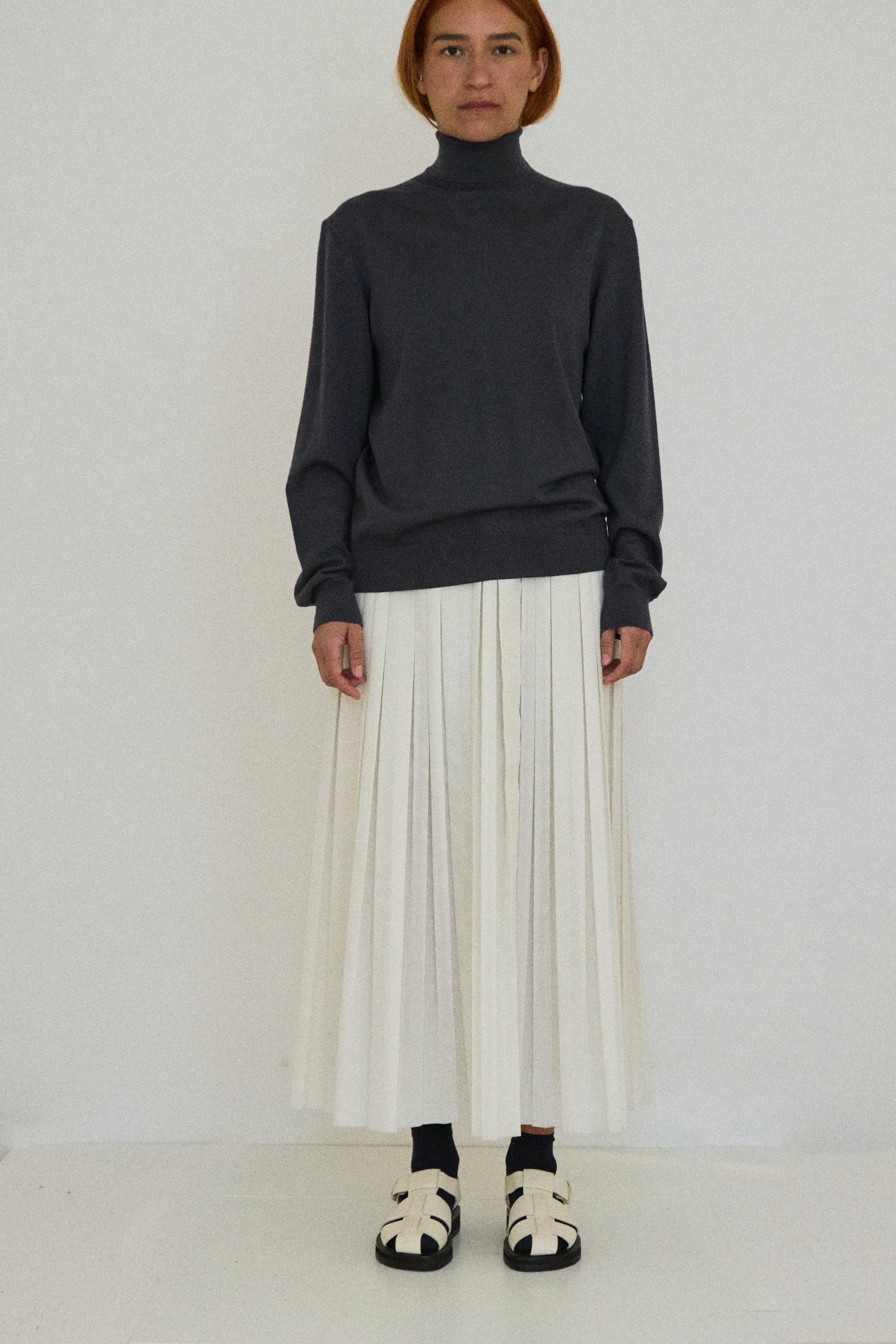 Person wearing a dark turtleneck sweater and white pleated skirt on a white background