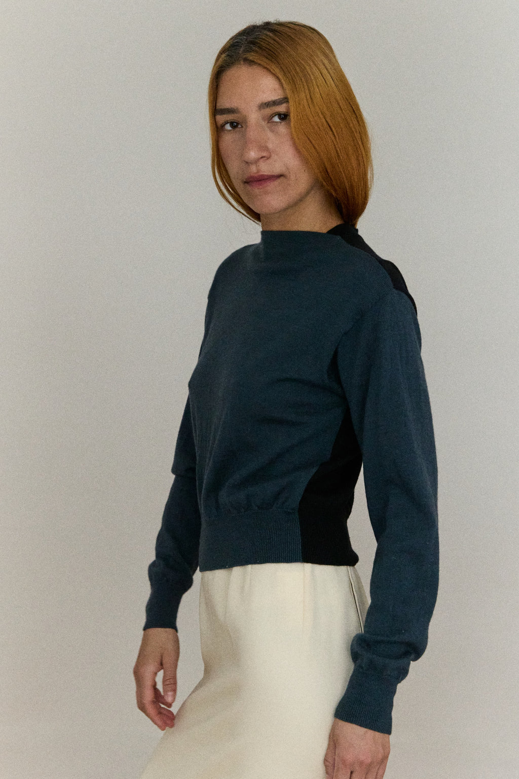 Woman wearing a dark blue turtleneck sweater and white pants against a plain background