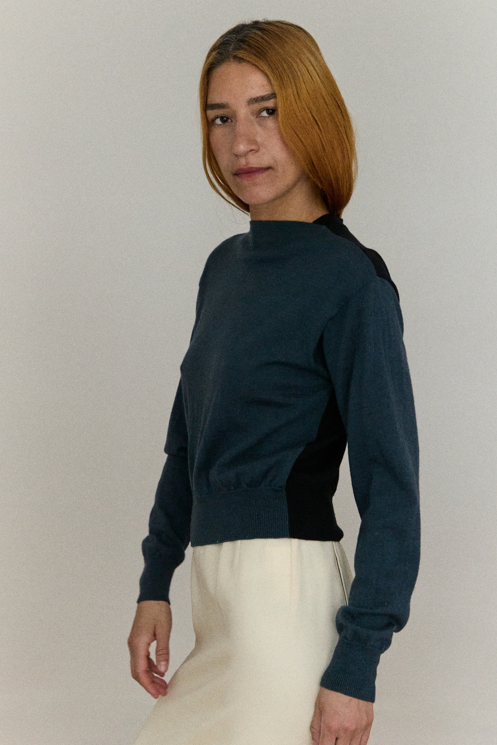 Woman wearing a dark blue turtleneck sweater and white pants against a plain background