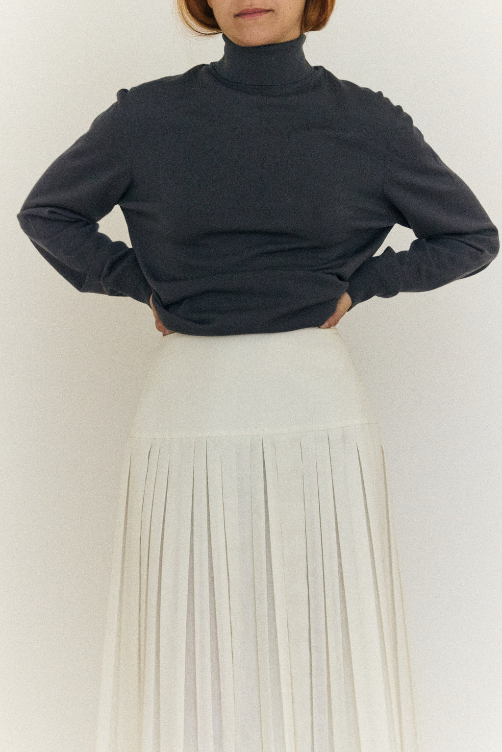 Person wearing a dark turtleneck sweater and light-colored pleated skirt against a white background