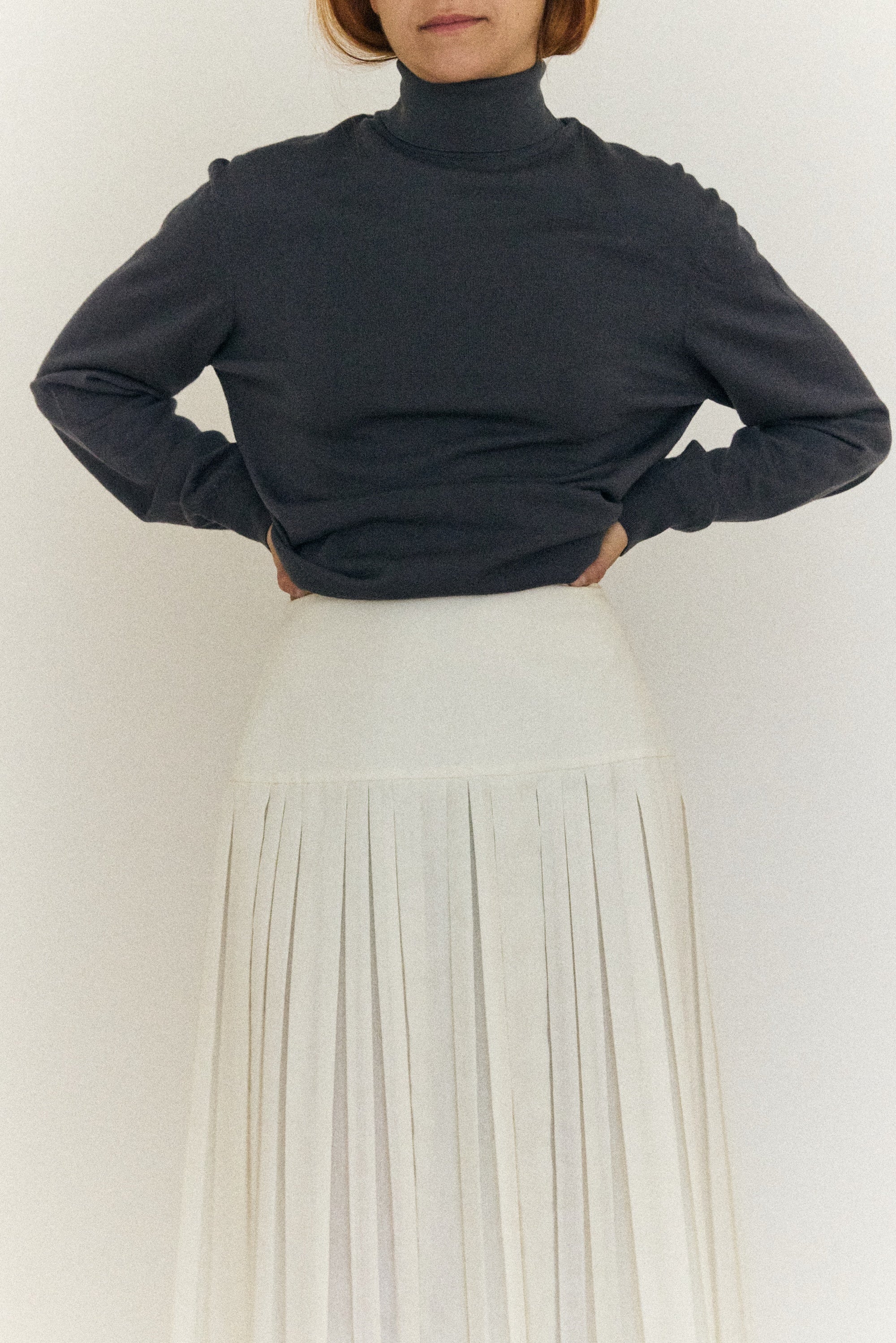 Person wearing a dark turtleneck sweater and light-colored pleated skirt against a white background