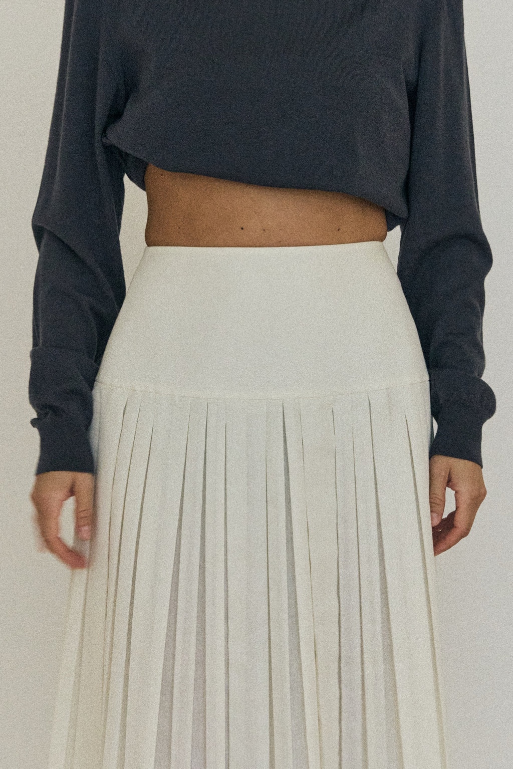 Person wearing a dark long-sleeve top and light pleated skirt on a plain background