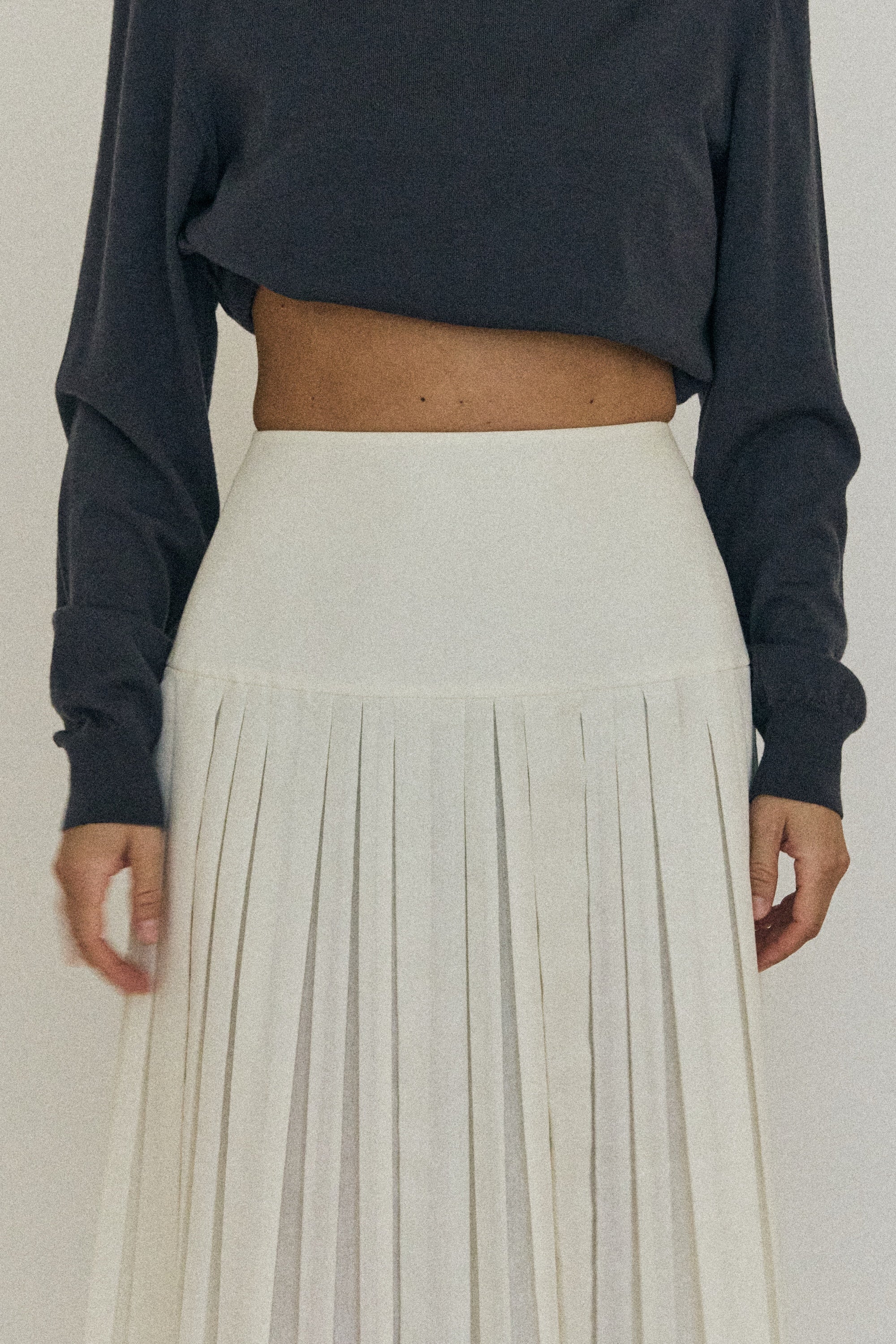 Person wearing a dark long-sleeve top and light pleated skirt on a plain background
