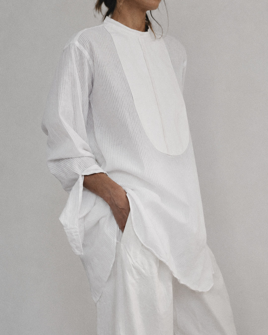 A person wearing a loose white long-sleeve top with vertical textured pleats and matching white wide-leg pants, photographed against a plain light gray studio background.
