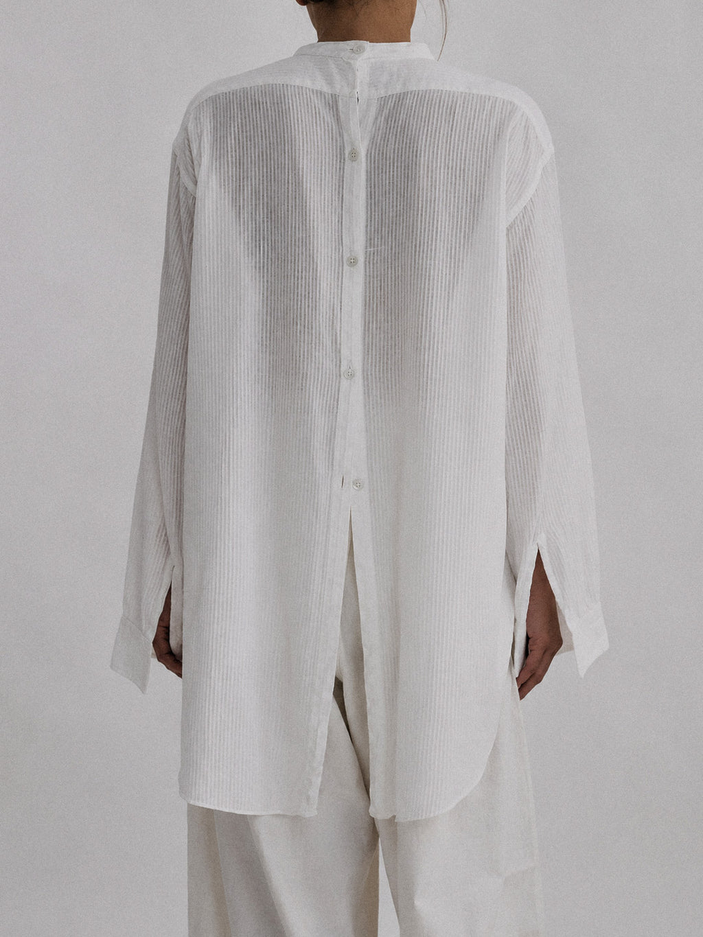 A person shown from the back wearing a loose white long-sleeve button-front top with a high collar and matching white pants against a plain light gray studio background.