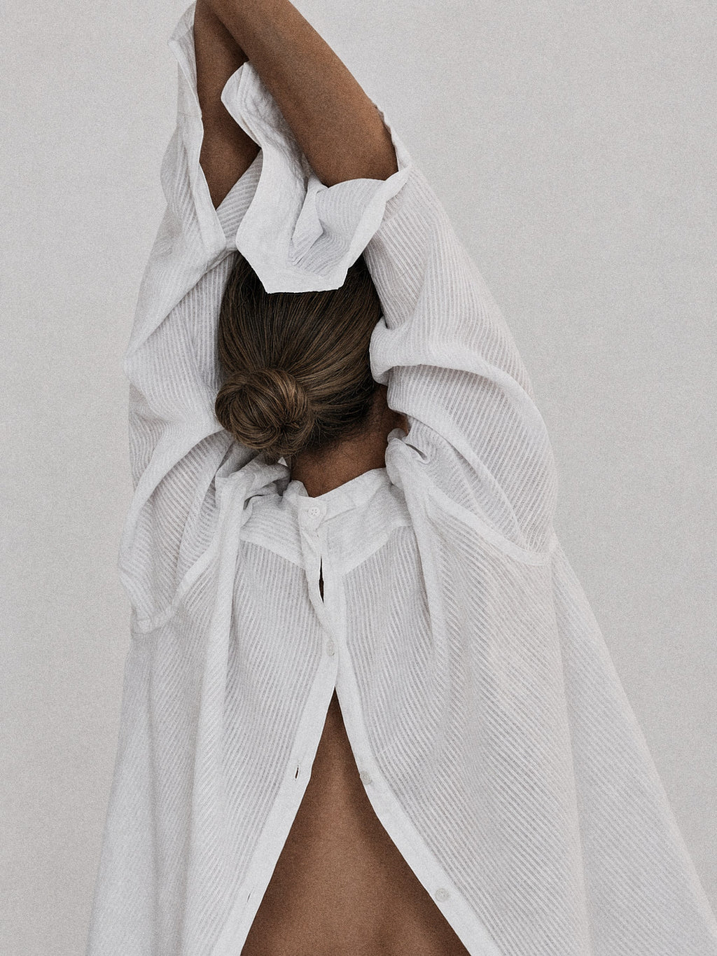 A close-up of a person wearing a loose white textured shirt with dramatic draped sleeves and a high open back, photographed against a light neutral background.