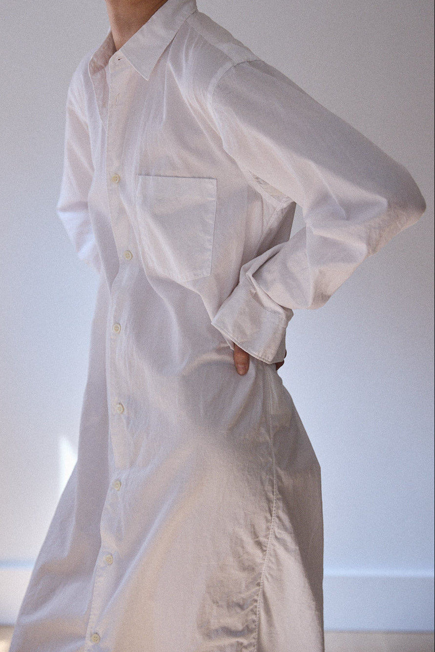 A woman wearing a loose white long-sleeve button-front shirt dress, standing against a plain light gray wall.