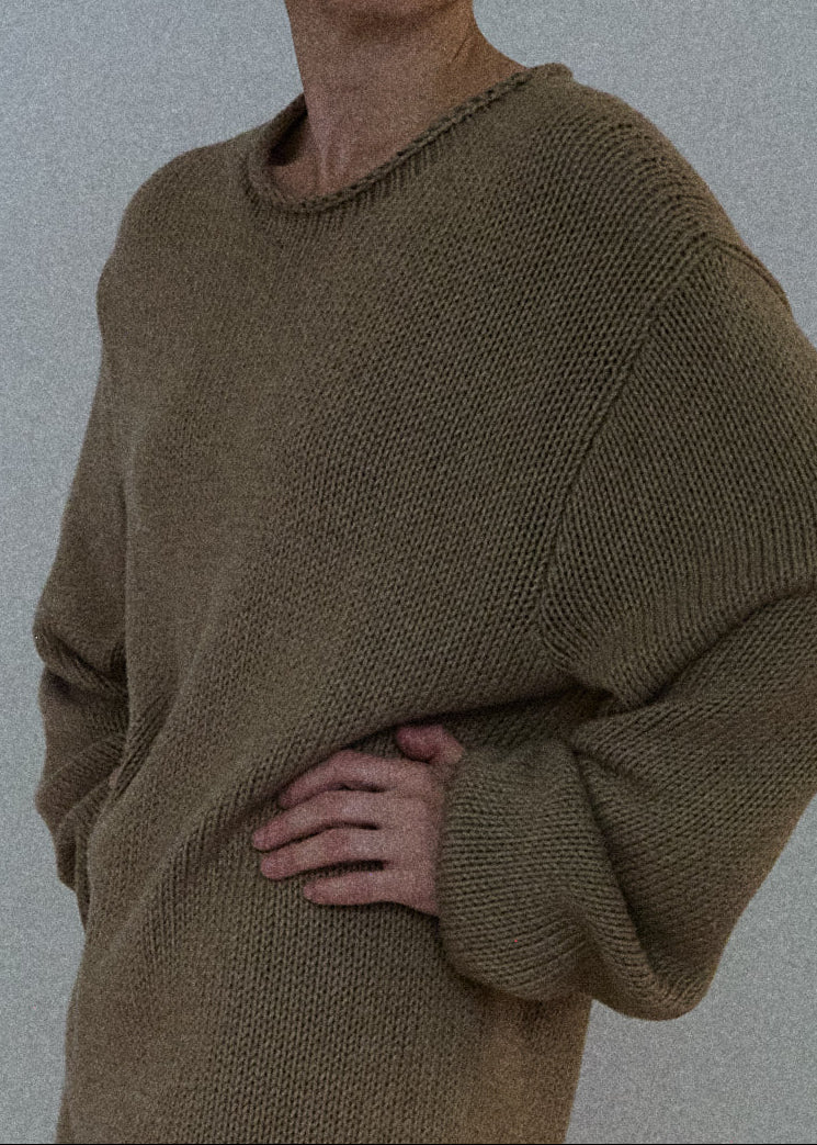 A person wearing an oversized ribbed knit sweater in a warm taupe-brown color, paired with blue jeans, shown against a plain light gray wall.