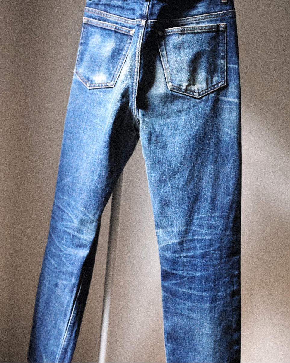 Blue denim jeans shown from the back, hanging from a white hanger against a plain beige wall.