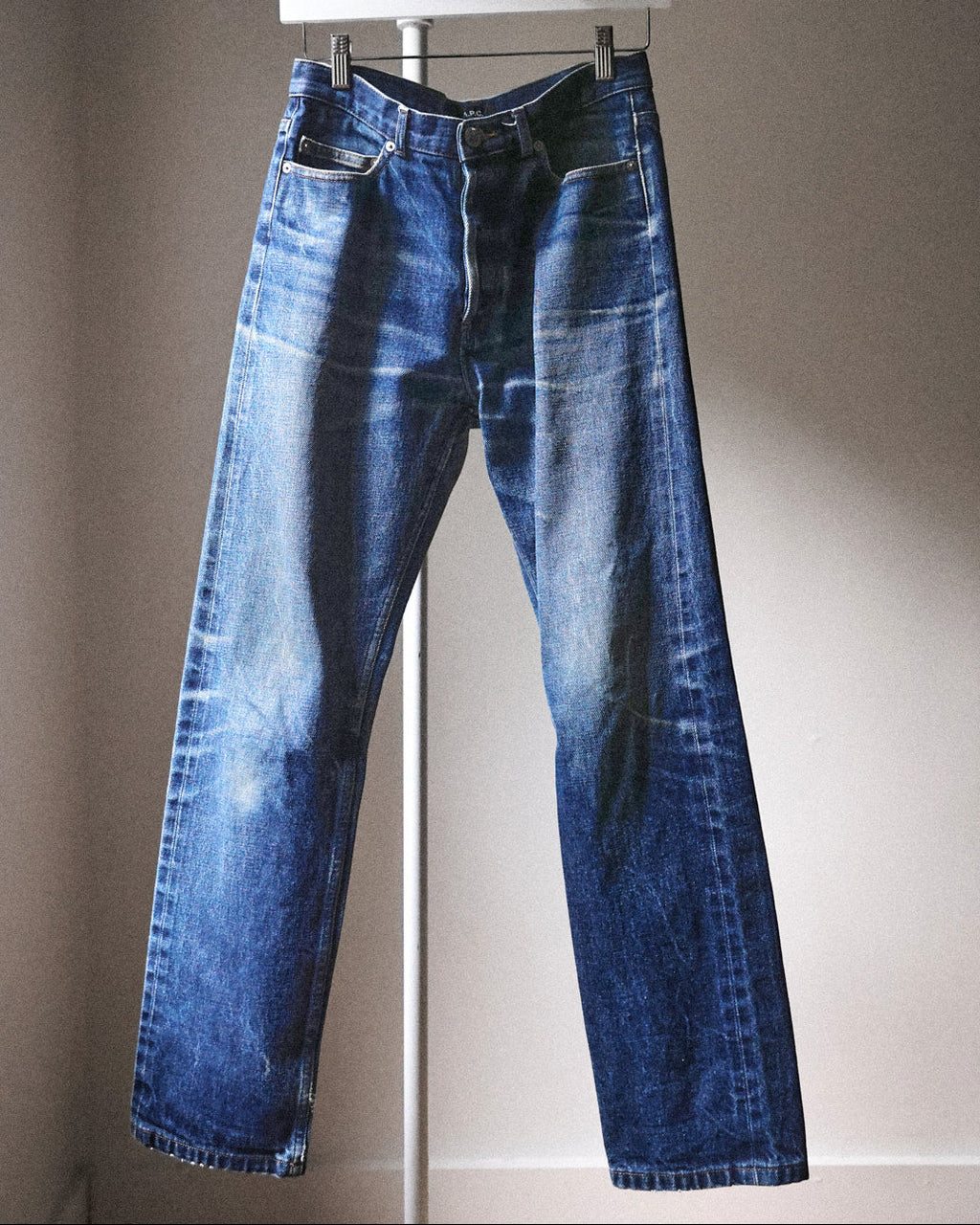 A pair of medium-wash blue denim jeans with a faded, distressed finish, hanging on a white hanger against a plain beige wall.
