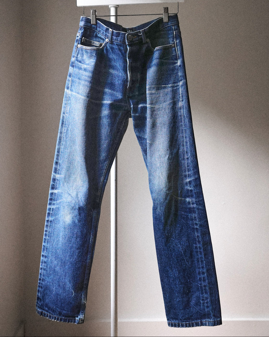 A pair of medium-wash blue denim jeans with a faded, distressed finish, hanging on a white hanger against a plain beige wall.