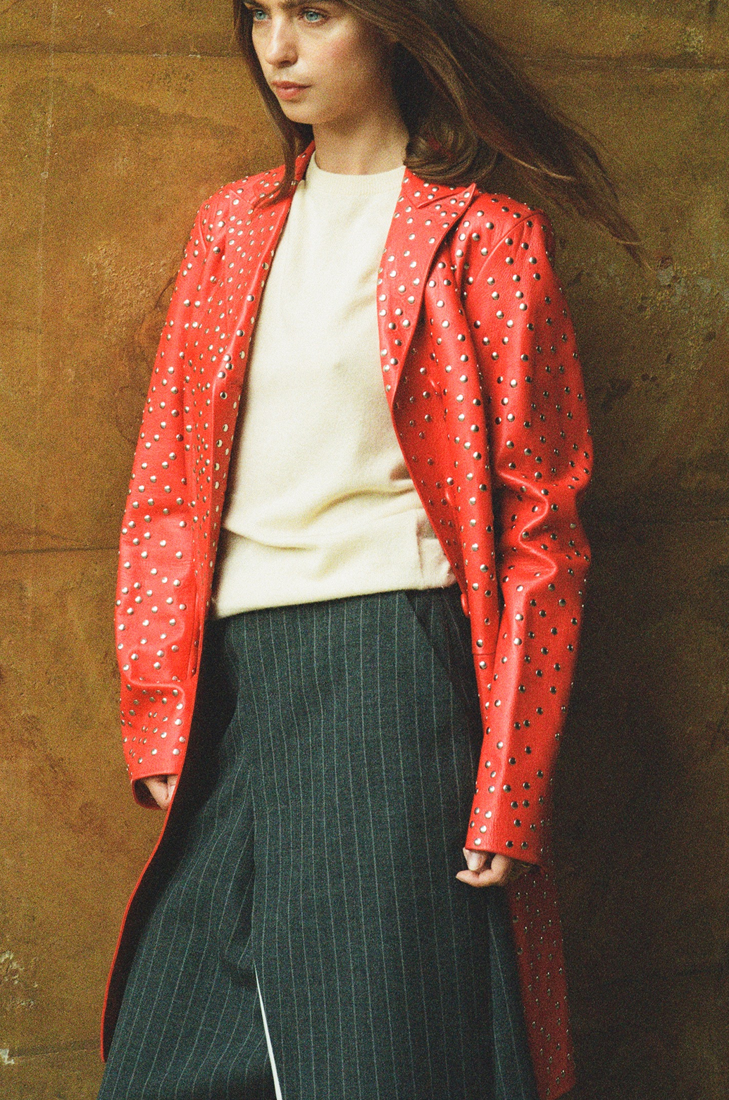 Woman wearing a red jacket with cut-out patterns, white top, and dark pinstripe pants against a textured wall.